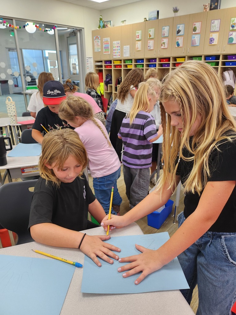 A high-angle view of elementary students collaborating at a desk. A blonde girl stands and helps trace another student's hand onto a piece of light blue paper using a yellow pencil. Classroom cubbies and word wall charts are visible in the background.