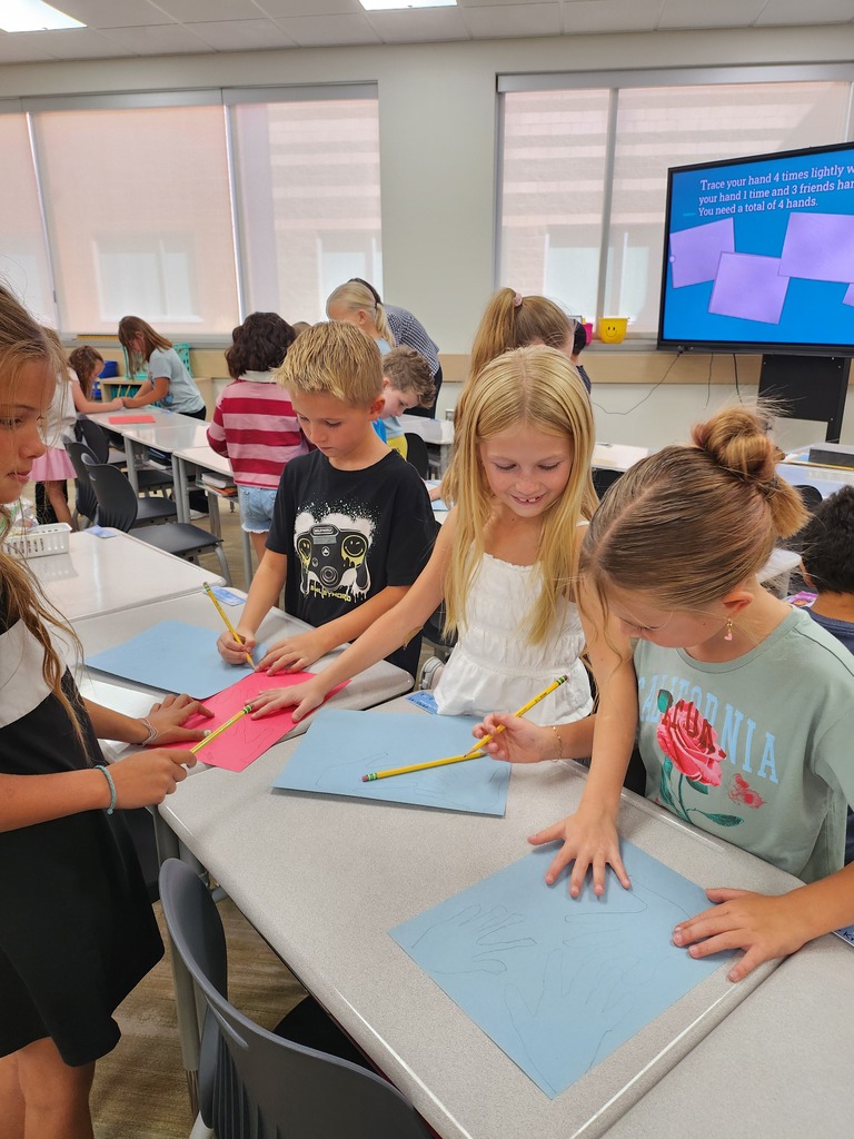 Students seated around classroom desks tracing their hands onto blue and red construction paper using pencils. The screen in the background shows instructions reading "Trace your hand 4 times lightly with pencil... You need a total of 4 hands." A girl in the foreground wears a shirt that says "CALIFORNIA."