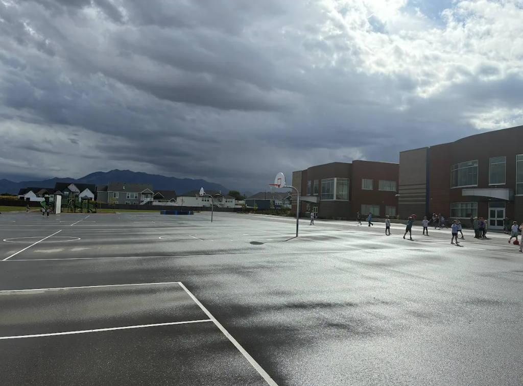 Wide shot of an outdoor asphalt school playground on a cloudy, overcast day. Children are scattered playing near the school building, with basketball hoops on the court, houses in the background, and mountains visible in the distance under a dark sky.