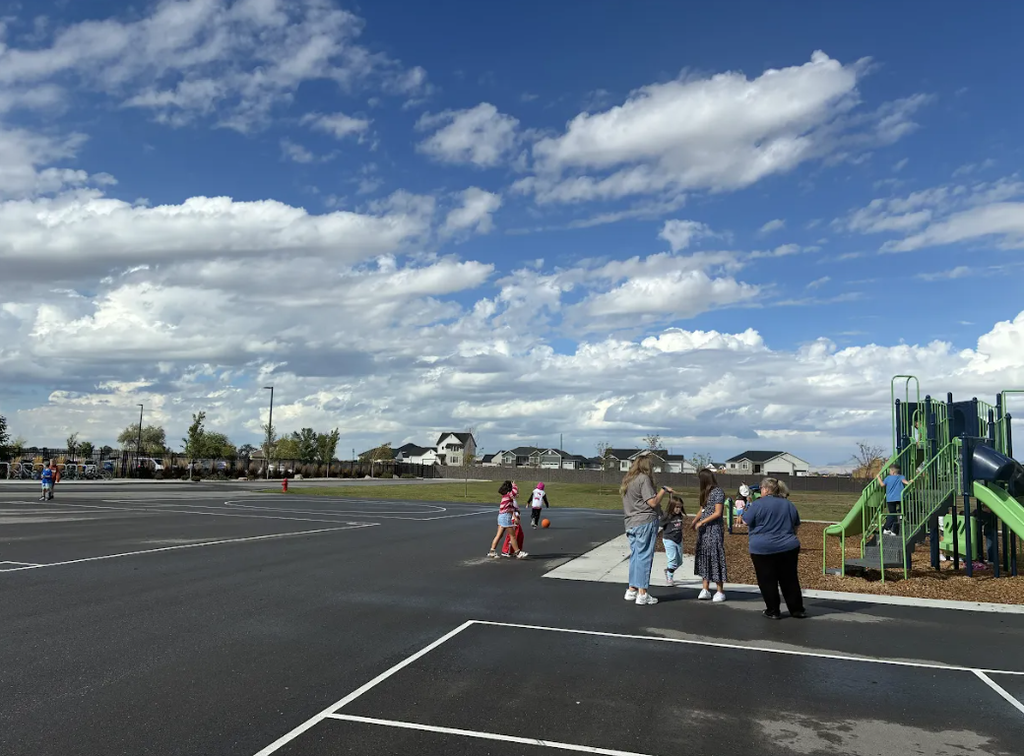 Children and adults standing and playing on a paved playground area next to a green playground structure under a bright, scattered-cloud blue sky, with suburban houses in the background.