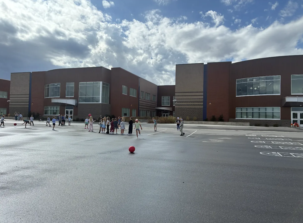 Children playing on an asphalt playground outside a modern brick school building under a partly cloudy sky, with a red dodgeball sitting in the middle ground near line painted games.