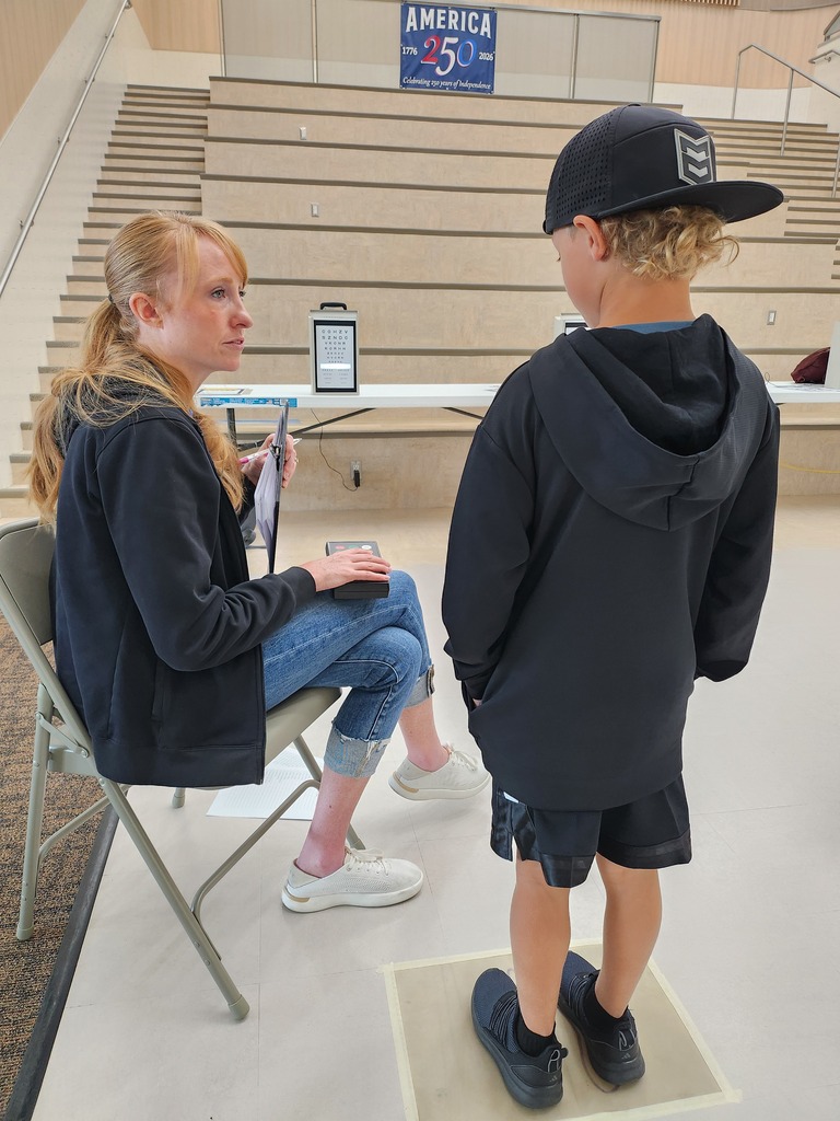 A young boy wearing a black baseball cap standing on a floor line while a woman in a black jacket and jeans conducts a vision screening with an eye chart set up in the background. A banner on the bleachers above reads "AMERICA 250 1776-2026: Celebrating 250 years of independence."