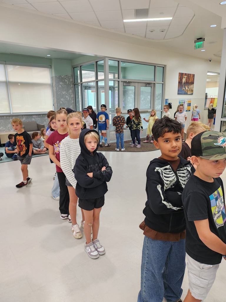 Elementary school students standing patiently in a line inside a brightly lit school hallway during a health screening day. In the background, an exit door sign reads "EXIT".