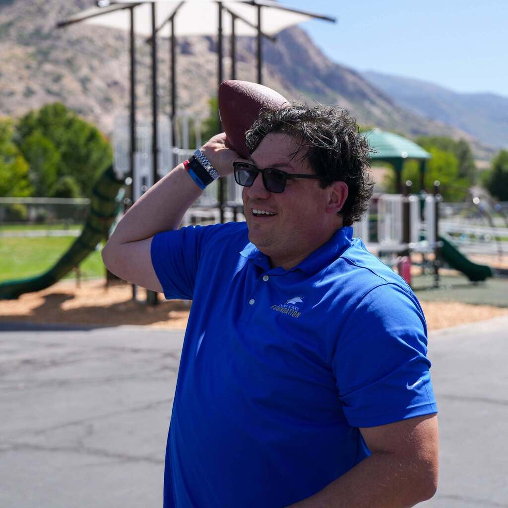 A district representative wearing sunglasses and a blue Foundation polo pulls back a football, getting ready to throw it on an outdoor playground with mountains visible in the background.