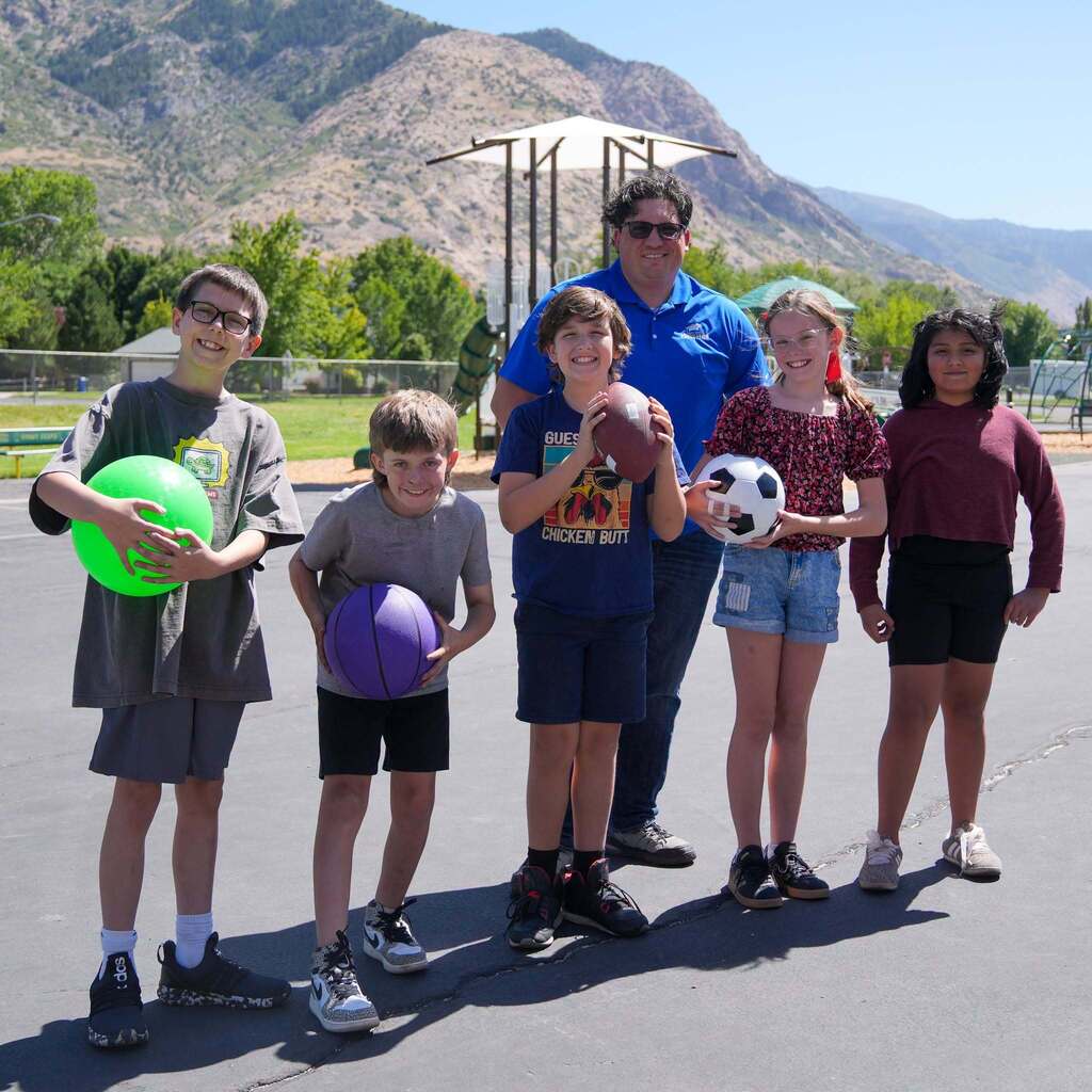 Five smiling elementary students stand outdoors on a paved playground holding various colorful sports balls, including a green playground ball, purple basketball, football, and soccer ball, alongside a adult district representative under a sunny mountain backdrop.