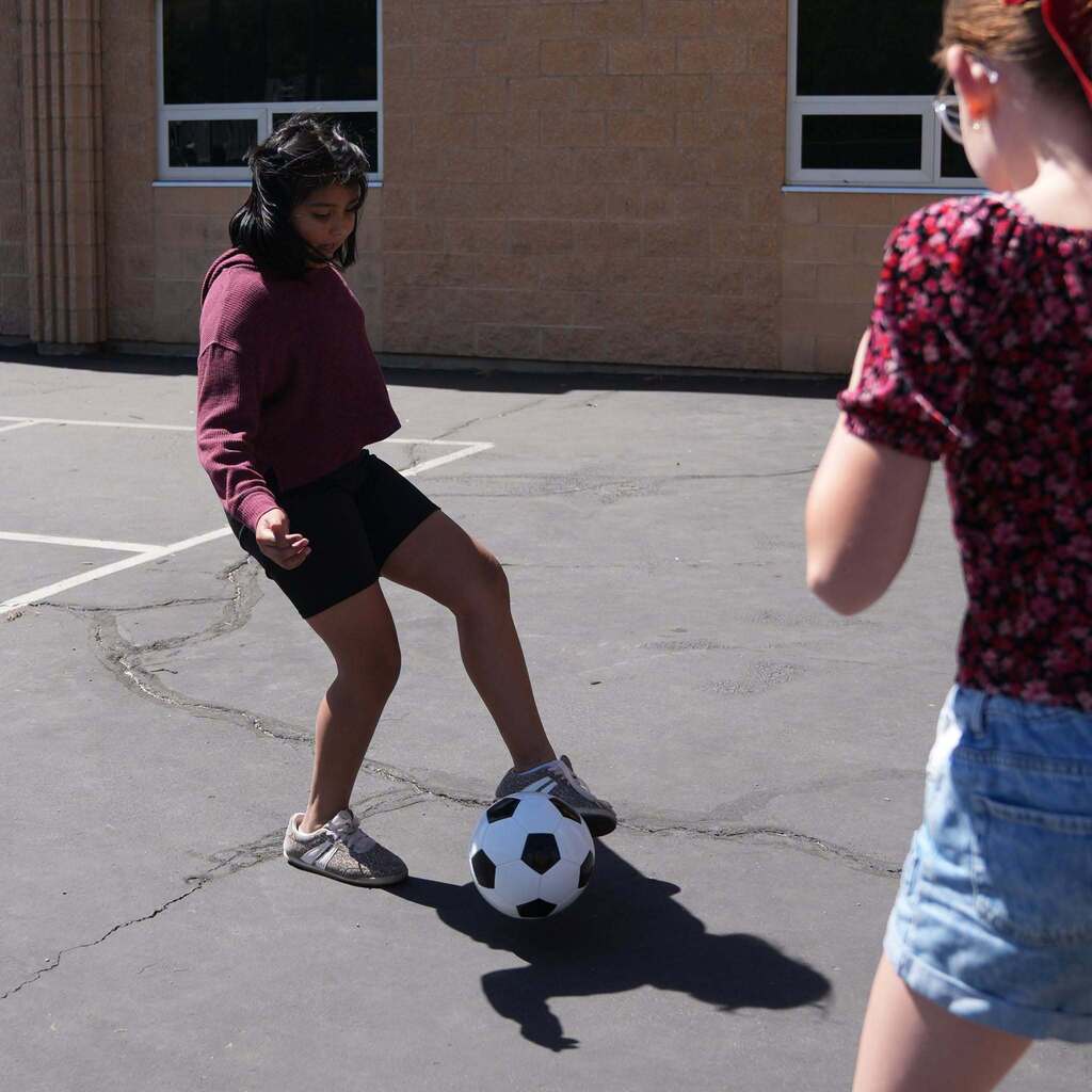 A female student outdoors on a blacktop schoolyard kicks a classic black-and-white soccer ball while another classmate looks on from the foreground.