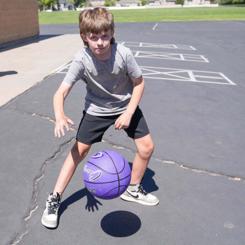 A young boy on an outdoor asphalt court focuses closely as he dribbles a bright purple basketball with one hand.