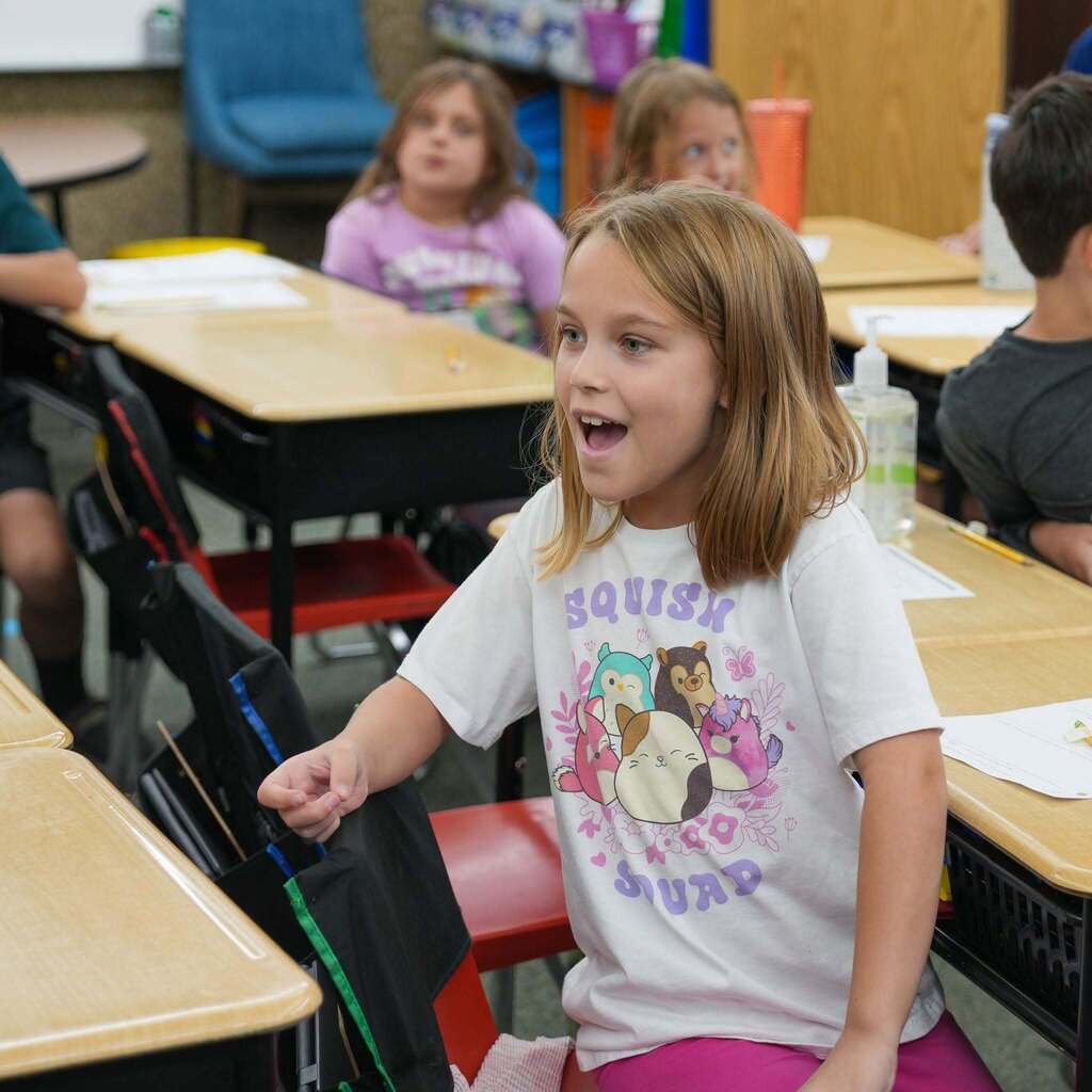 A young female student wearing a white shirt with a colorful graphic reacts with excitement, smiling open-mouthed as she leans forward at her classroom desk during a presentation.