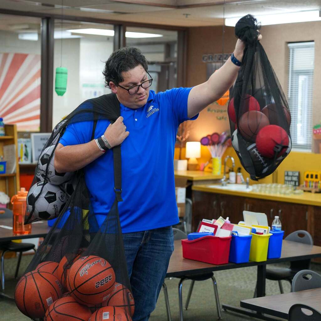 A Weber School District Foundation representative wearing a blue polo shirt and glasses stands inside a classroom, holding up a black mesh equipment bag filled with red dodgeballs and footballs while carrying large mesh bags full of basketballs and soccer balls over his shoulder.