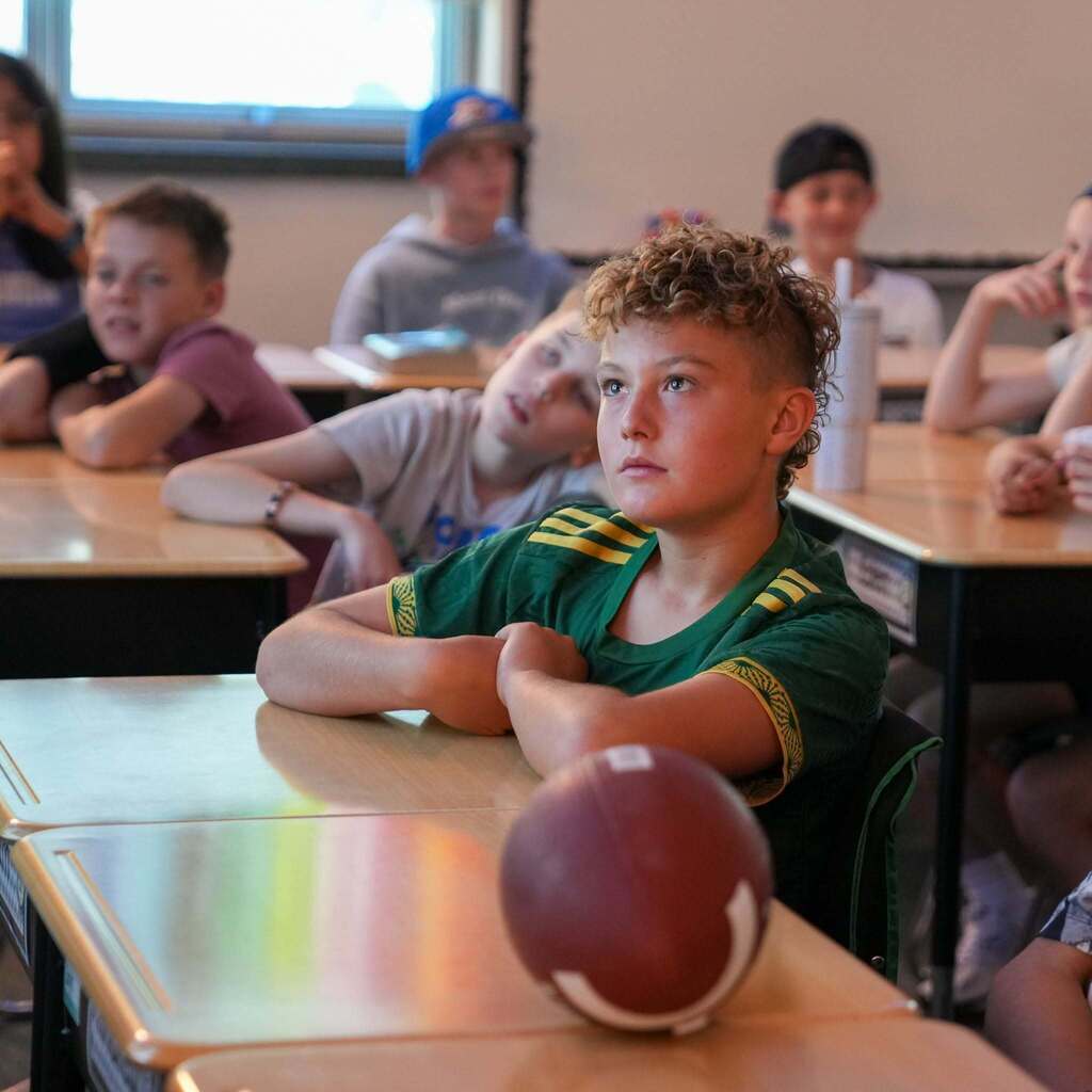 A male student with curly hair sits attentively at his desk with his arms crossed, focusing on a lesson while a miniature football rests on the desk in front of him.