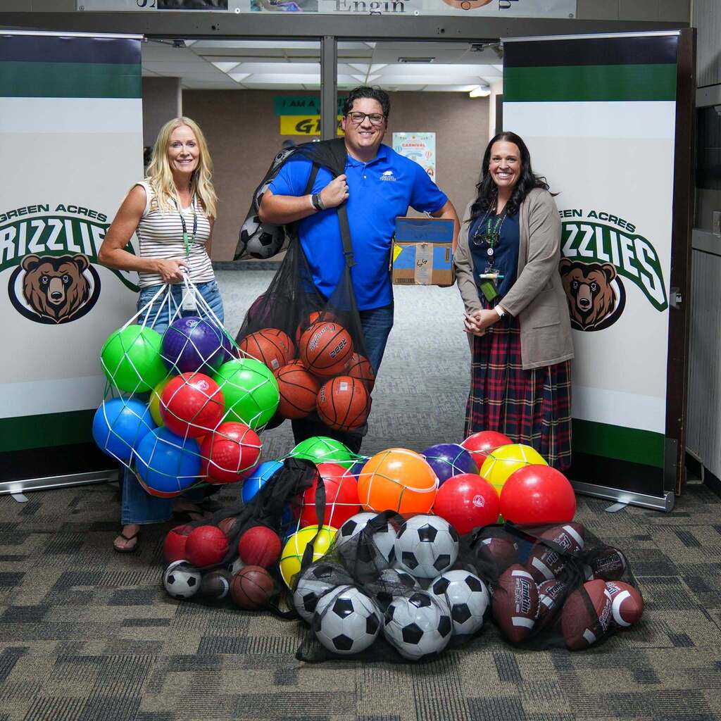 Three adults pose indoors at Green Acres Elementary surrounded by dynamic sports equipment. A man in a blue Weber School District Foundation polo shirt stands in the center holding mesh bags of basketballs and soccer balls, flanked by two smiling women holding colorful playground balls, with Green Acres Grizzlies banners in the background.