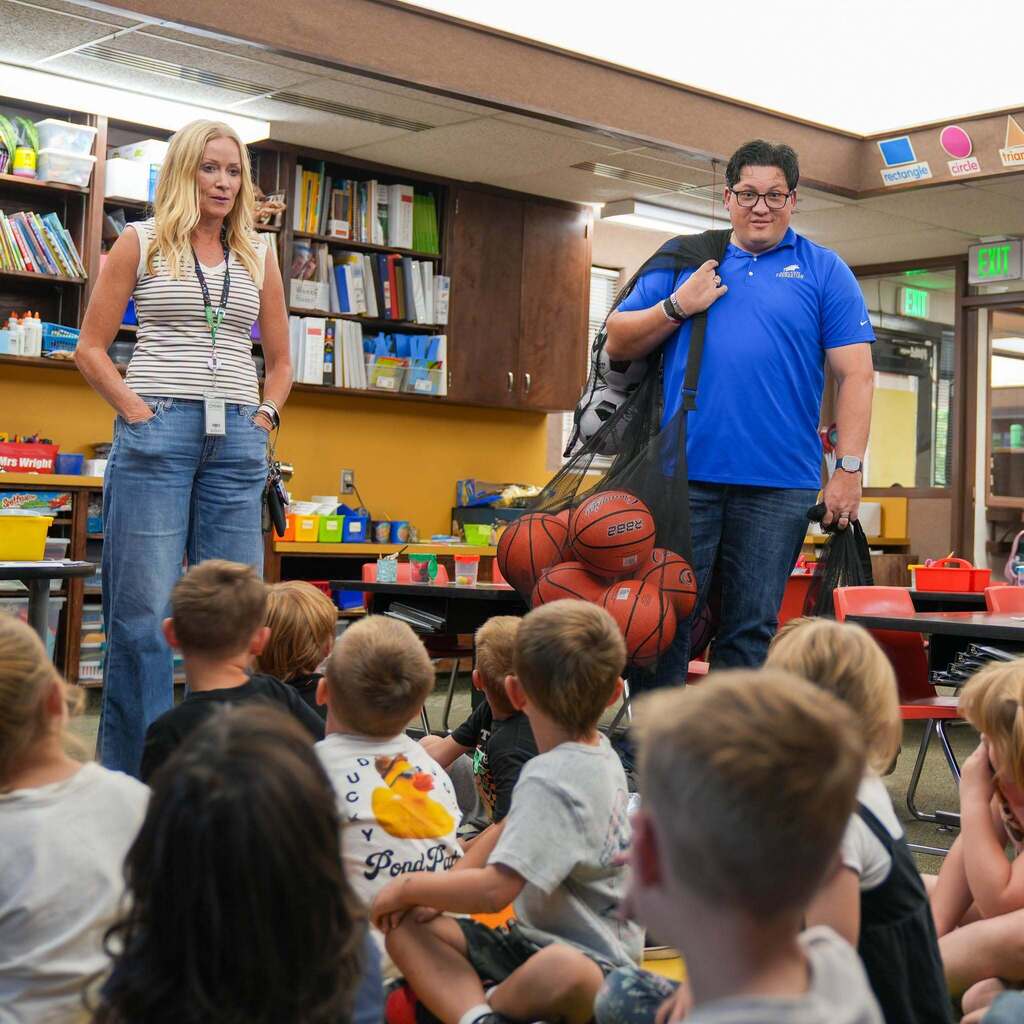 A board member and a district representative holding a large mesh bag filled with basketballs present to a classroom full of elementary students sitting attentively on the floor.