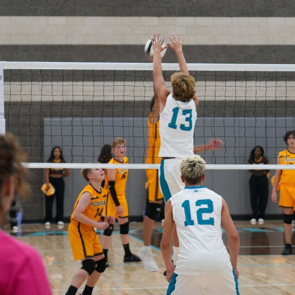 A volleyball player wearing number 13 in a white jersey leaps at the net with arms fully extended to execute a two-handed block against an incoming attack, with teammates positioning behind him.