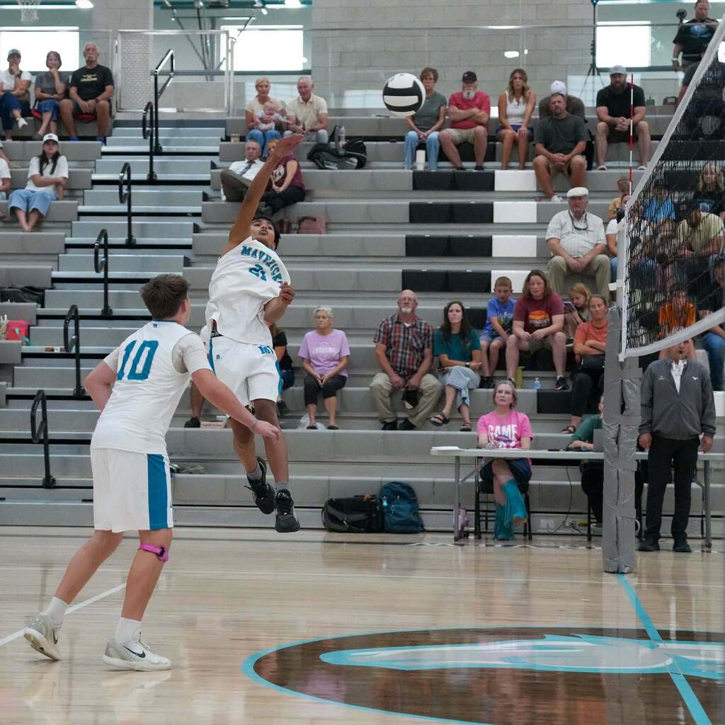 A Mavericks volleyball player wearing number 21 leaps high for a powerful spike over the net while a teammate wearing number 10 watches the play from the front court.