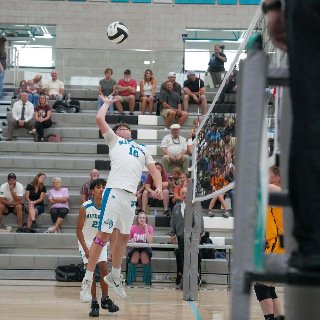 A male volleyball player wearing a white Mavericks uniform with the number 10 leaps high near the net to serve or spike a volleyball during an indoor middle school match, with spectators watching from the bleachers.
