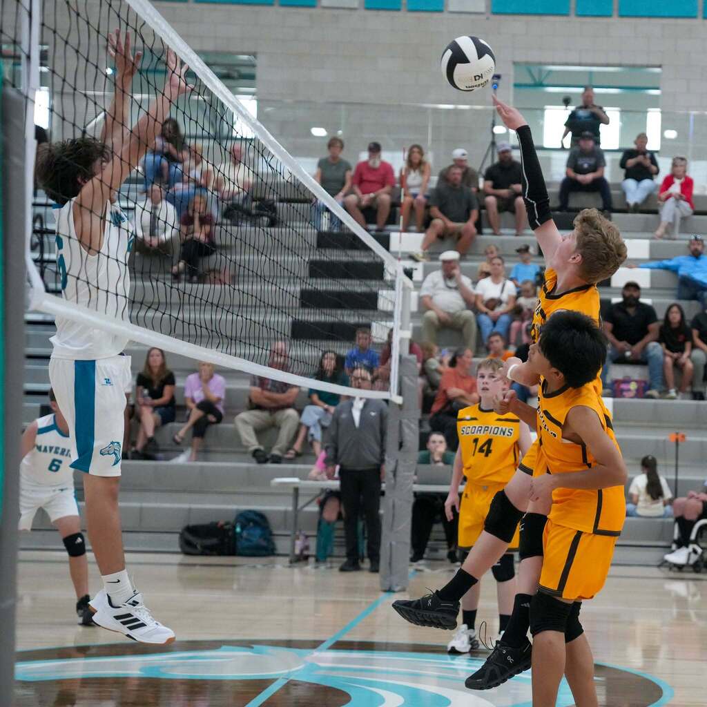 A Scorpions player jumps to tip the volleyball over the net while an opposing Mavericks player reaches high to contest the shot at the net.