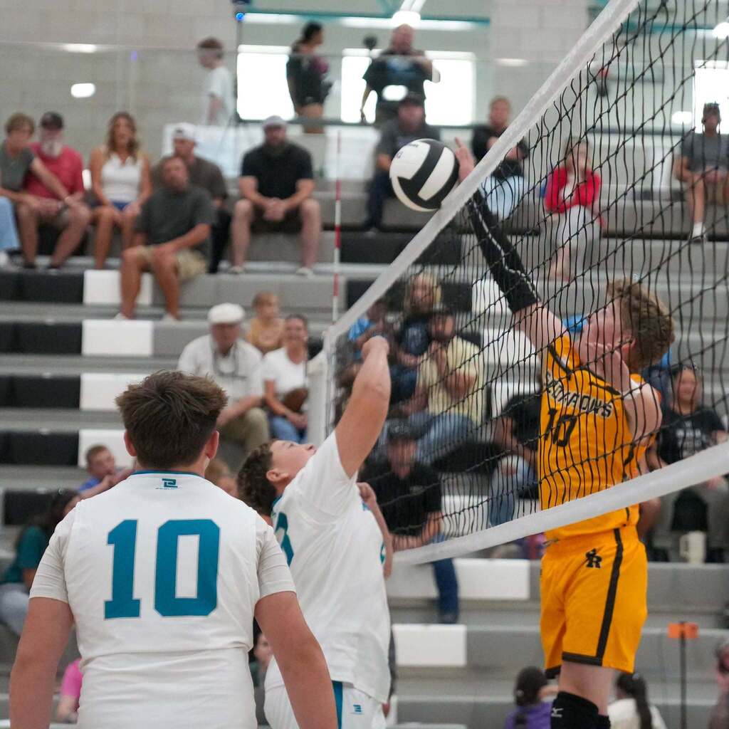 A Scorpions volleyball player wearing jersey number 10 reaches high over the net to block or spike the ball while Mavericks players prepare to defend below.