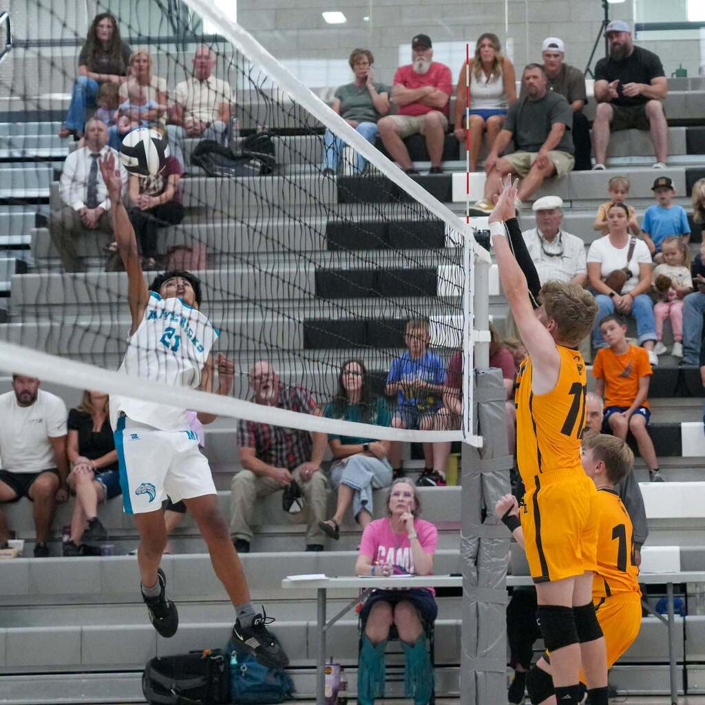 A Mavericks volleyball player wearing a white jersey with the number 21 jumps above the net to spike the ball while two Scorpions players in yellow jerseys jump up to attempt a block.