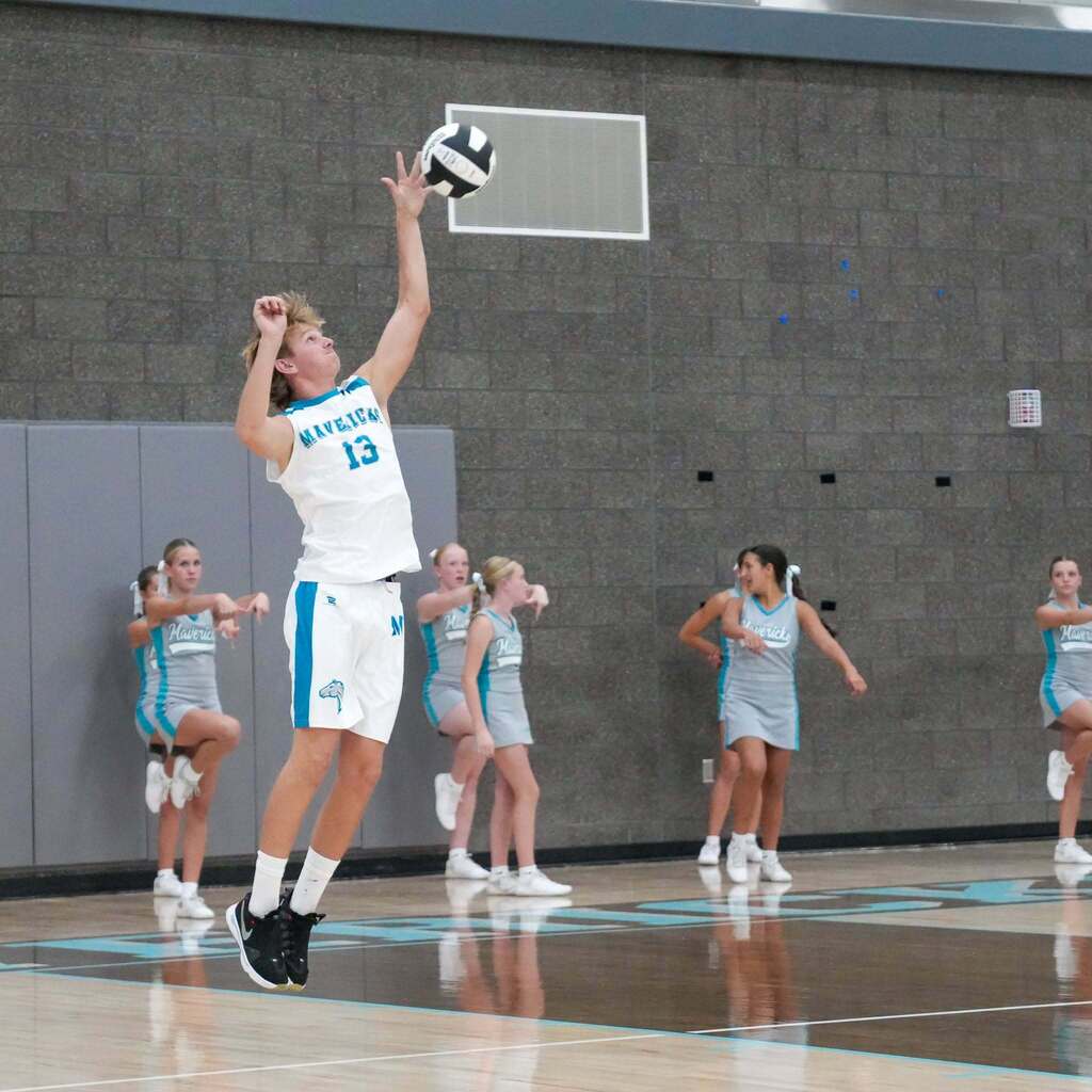 A male student-athlete wearing a white Mavericks uniform jumps high into the air to serve a volleyball on a polished gymnasium floor while cheerleaders perform along the baseline.