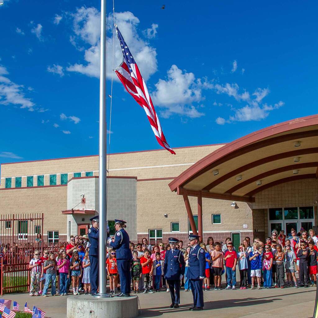 Honor Guard members raise a fresh American flag up a tall flagpole outside West Haven Elementary while students and staff stand below with hands over their hearts against a bright blue sky.