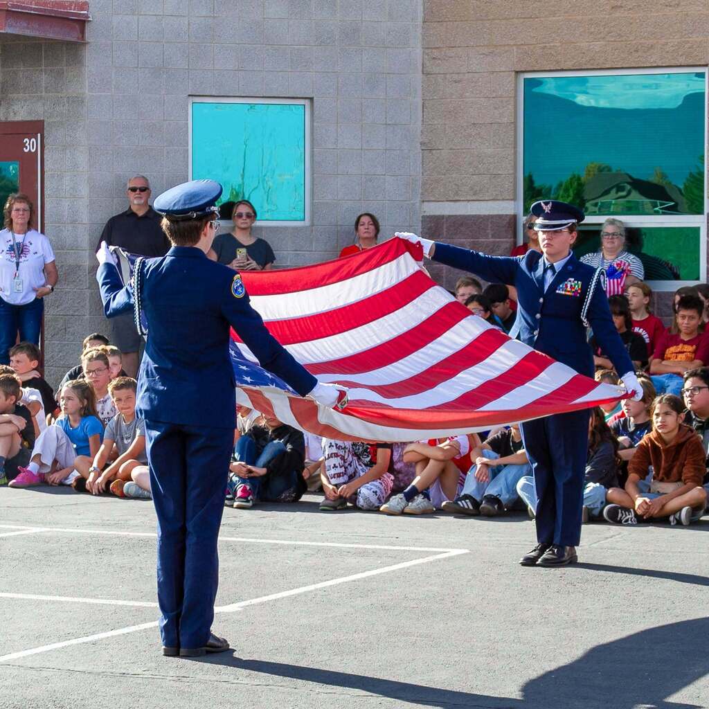 Two members of the Utah Military Academy Honor Guard dressed in blue dress uniforms hold a large American flag outstretched above the pavement while Elementary students sit in the background watching respectfully.