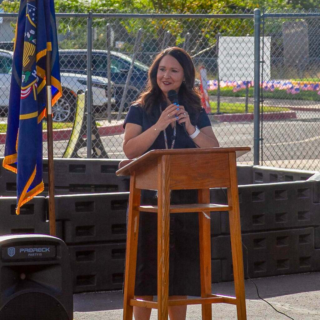 Principal Courtney Geisler speaks into a microphone at a wooden podium outdoors, addressing students and community guests near a Utah state flag during the West Haven Elementary flag retirement ceremony.
