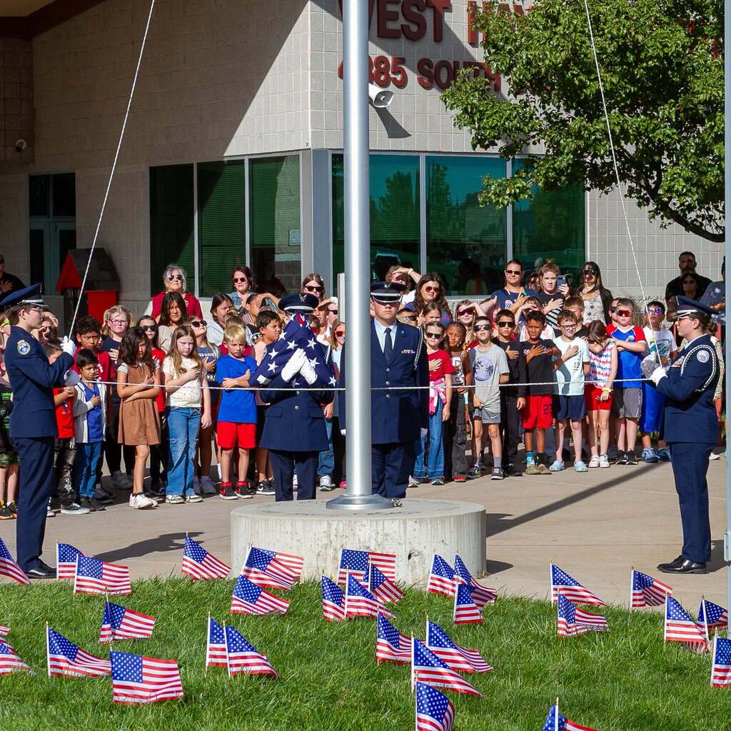 Four Honor Guard members stand at attention near a flagpole surrounded by a field of small American flags stuck in the lawn, while audience members stand behind them with hands on their hearts.