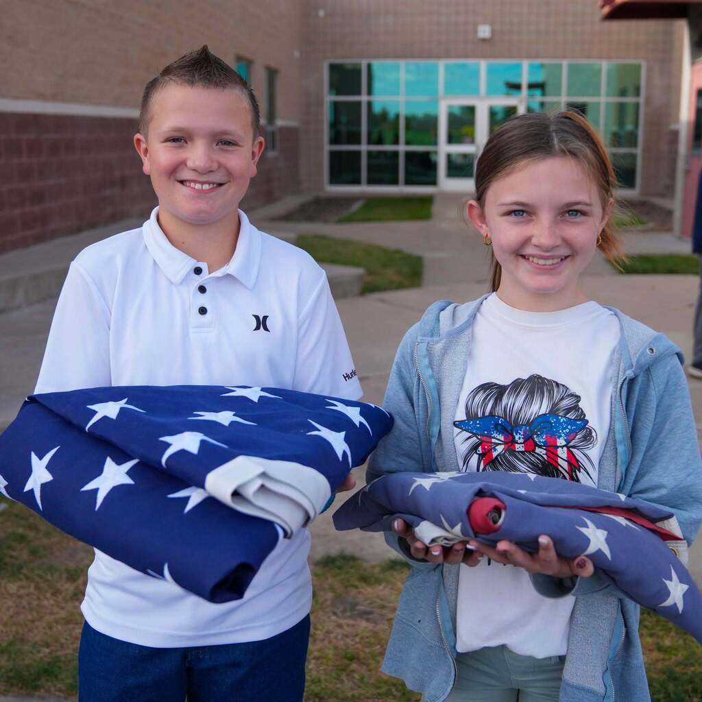 Two smiling West Haven Elementary students stand side by side outside their school, each holding a neatly folded American flag presented during the ceremony.