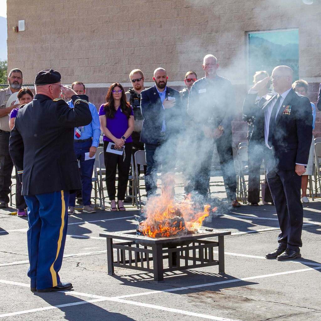 A military official in uniform salutes across a burning fire pit where worn flags are being respectfully retired, as community leaders, district administrators, and guests look on solemn-faced.