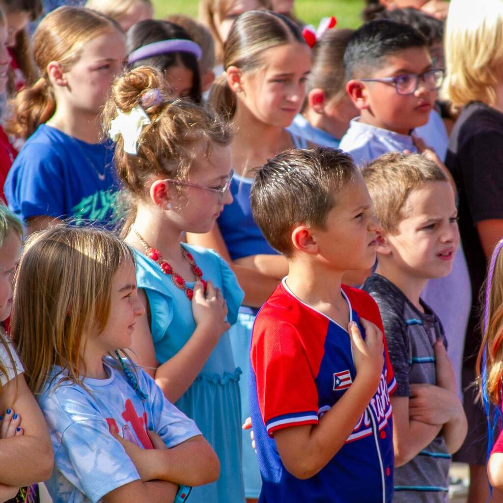 A close-up view of elementary school students standing outdoors in a row, each placing a hand over their heart with focused expressions during the patriotic flag ceremony.
