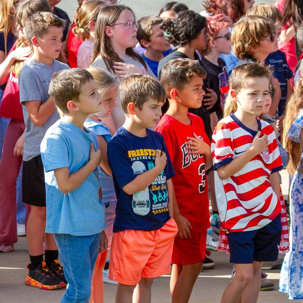 A line of young Hawk students dressed in patriotic red, white, and blue clothing stand attentive in a crowd with their right hands placed over their hearts.