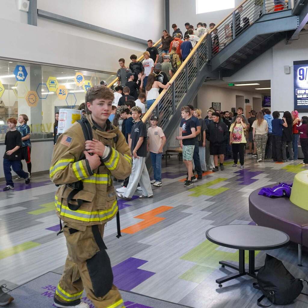 A student wearing full firefighter turn-out gear walks through a bustling school hallway during Innovation High's remembrance stair climb.