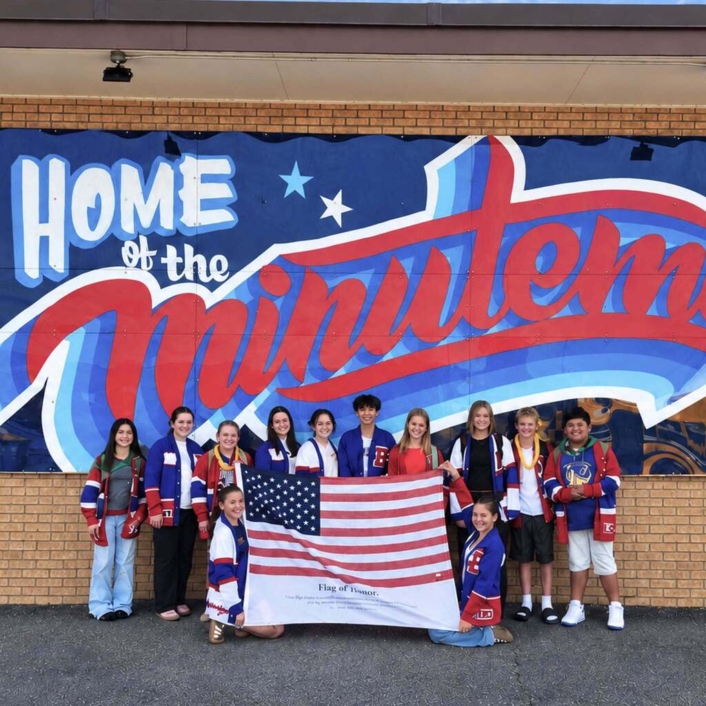 Junior high students line up outdoors in front of a blue and red "Home of the Minutemen" mural while holding up an American Flag of Honor.