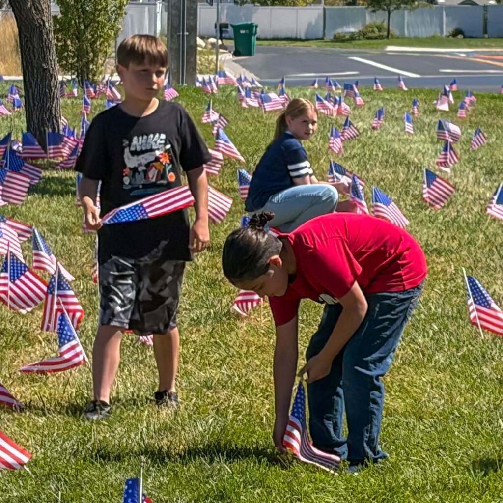 Elementary students bend down to carefully plant small American flags into the grass on their school lawn as part of a 9/11 Field of Flags tribute.