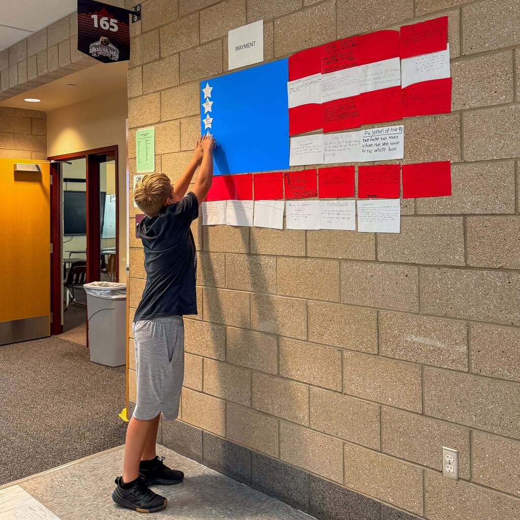 A student reaches up to affix a patriotic paper star to a large, hand-crafted American flag display posted on a brick hallway wall at Rocky Mountain Junior High.