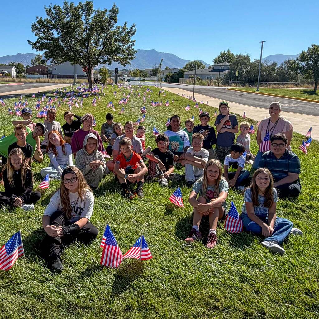 A group of West Haven Elementary students and staff pose happily while sitting outdoors in the green grass among hundreds of small American flags planted on the school lawn.