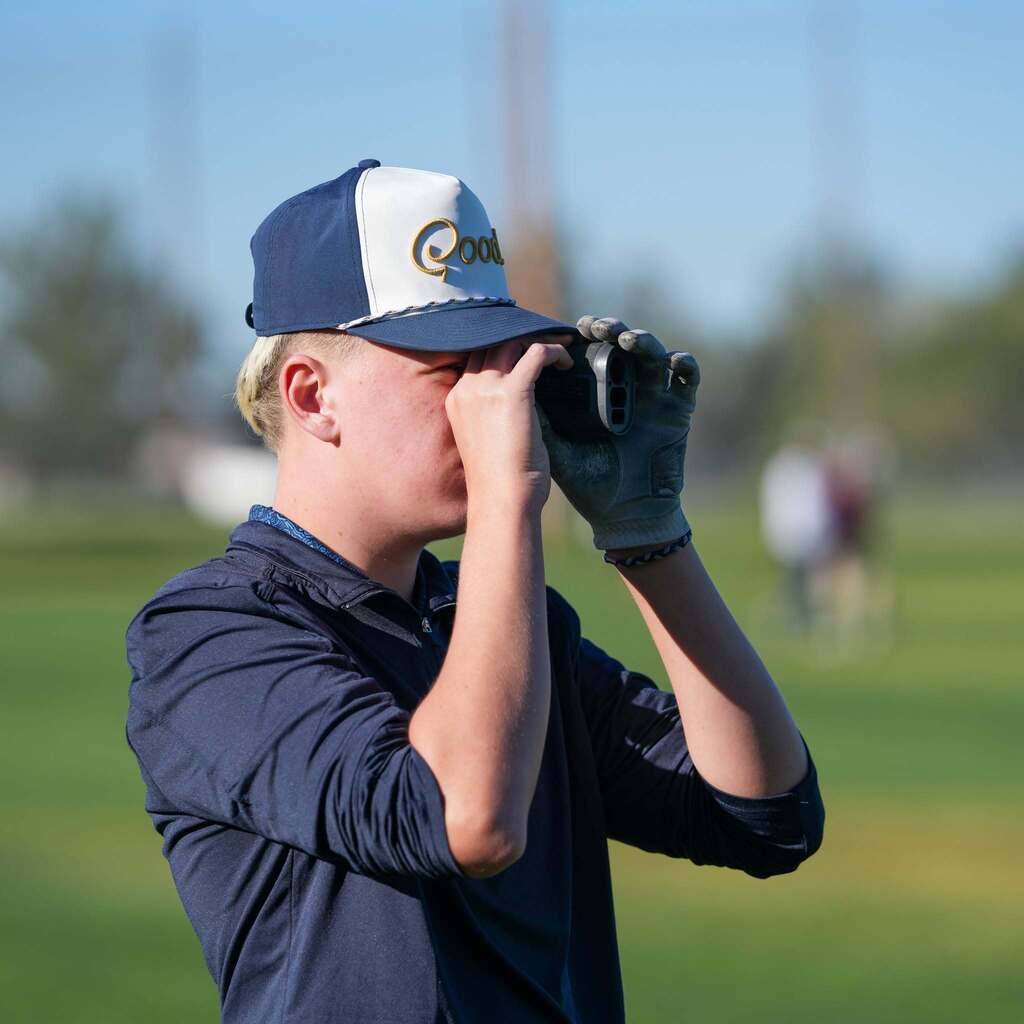 A close-up side profile of a male golfer wearing a navy and white cap, using a handheld digital rangefinder to measure distance on the course.
