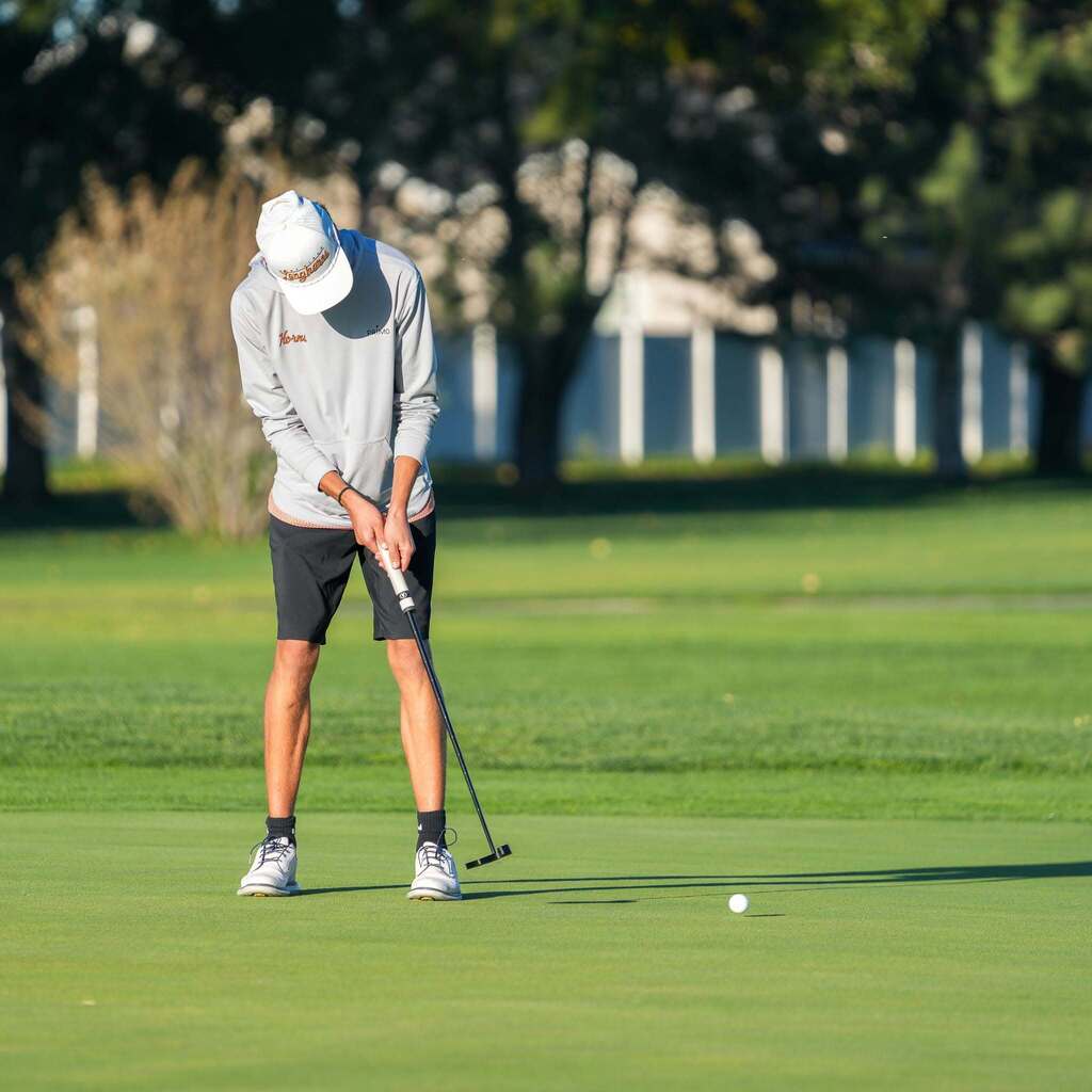 A high school golfer bends forward at the waist on the putting green, maintaining focus as his putter sends the ball rolling smoothly toward the hole.