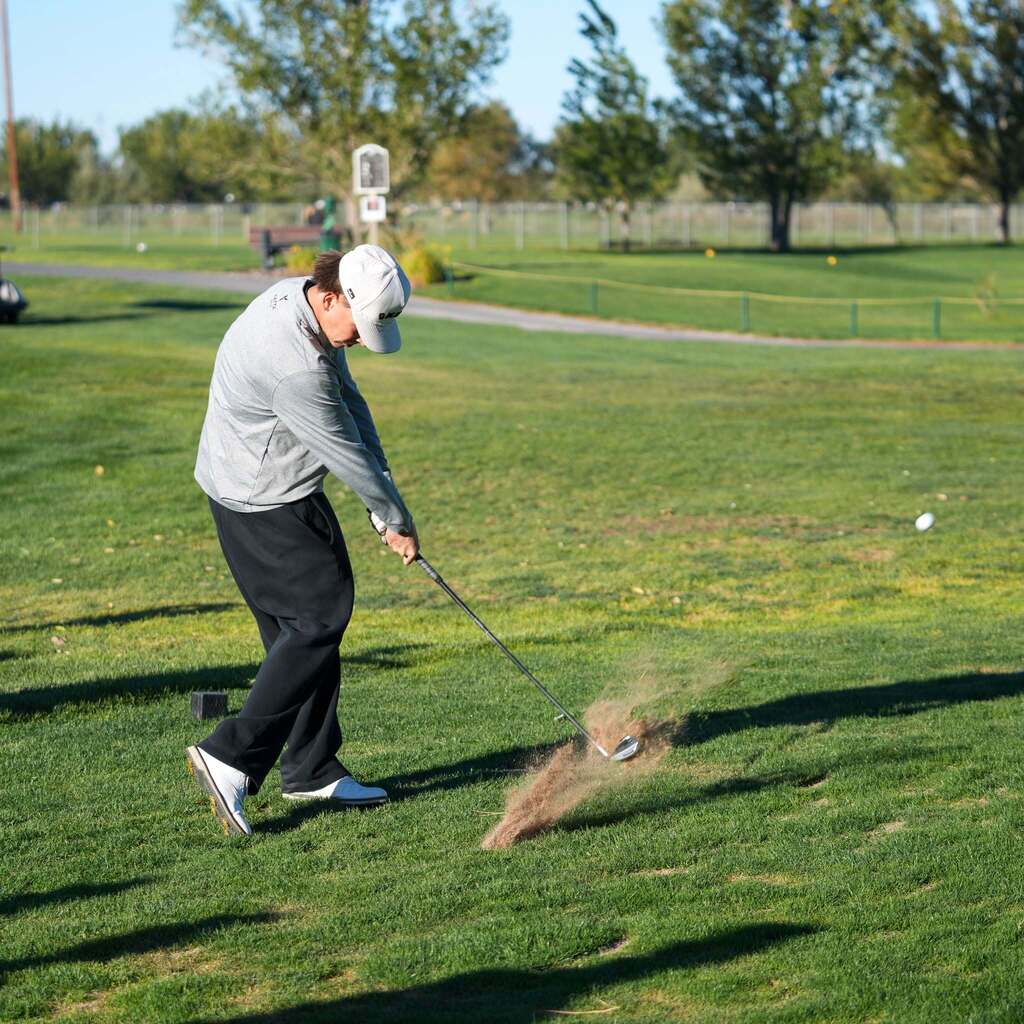 A close-up view of a high school golfer swinging through an iron shot on the fairway, creating a cloud of dust and grass divot as the club strikes the turf.