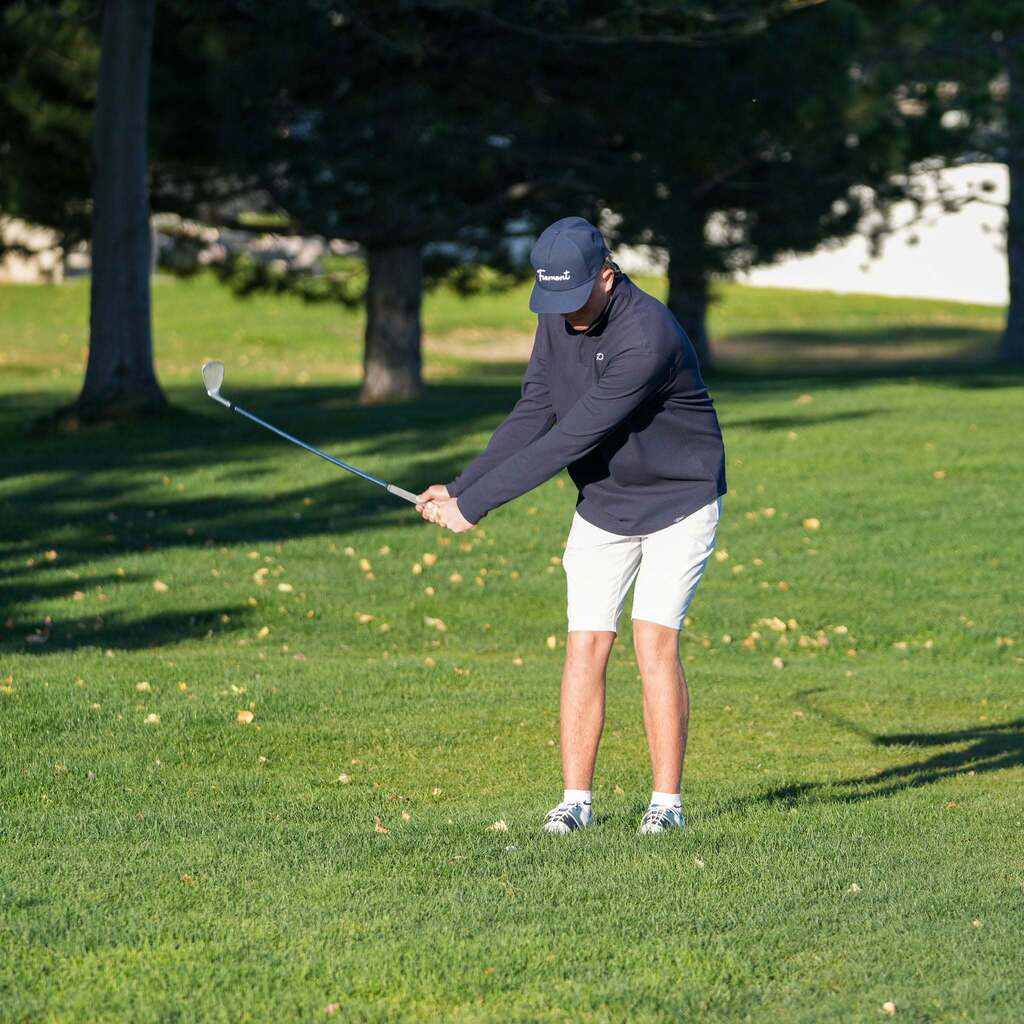 A Fremont High golfer dressed in a navy long-sleeve shirt and white shorts initiates his downswing with an iron from the fairway grass.