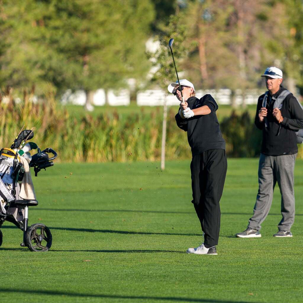 A high school golfer in a dark sweatshirt and white cap swings his club mid-pitch shot, while a teammate or coach stands nearby on the fairway watching intently.