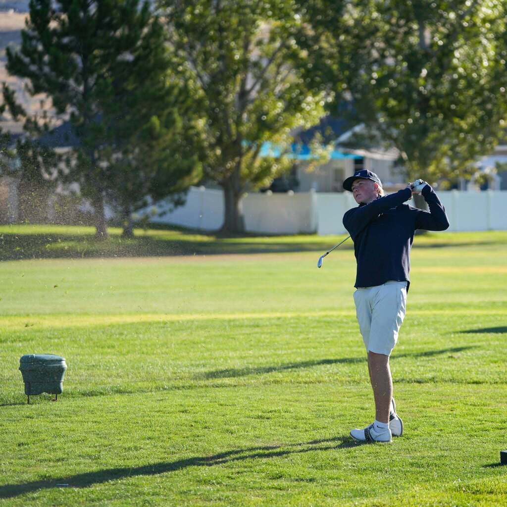 A high school golfer in a navy long-sleeve shirt and white shorts holds his follow-through stance after taking a swing off the tee box at Remuda Golf Course.