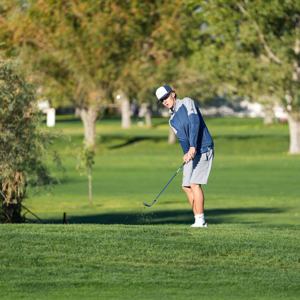 A golfer in a blue windbreaker jacket and grey shorts tracks his ball in mid-air right after launching a crisp iron shot off the turf.