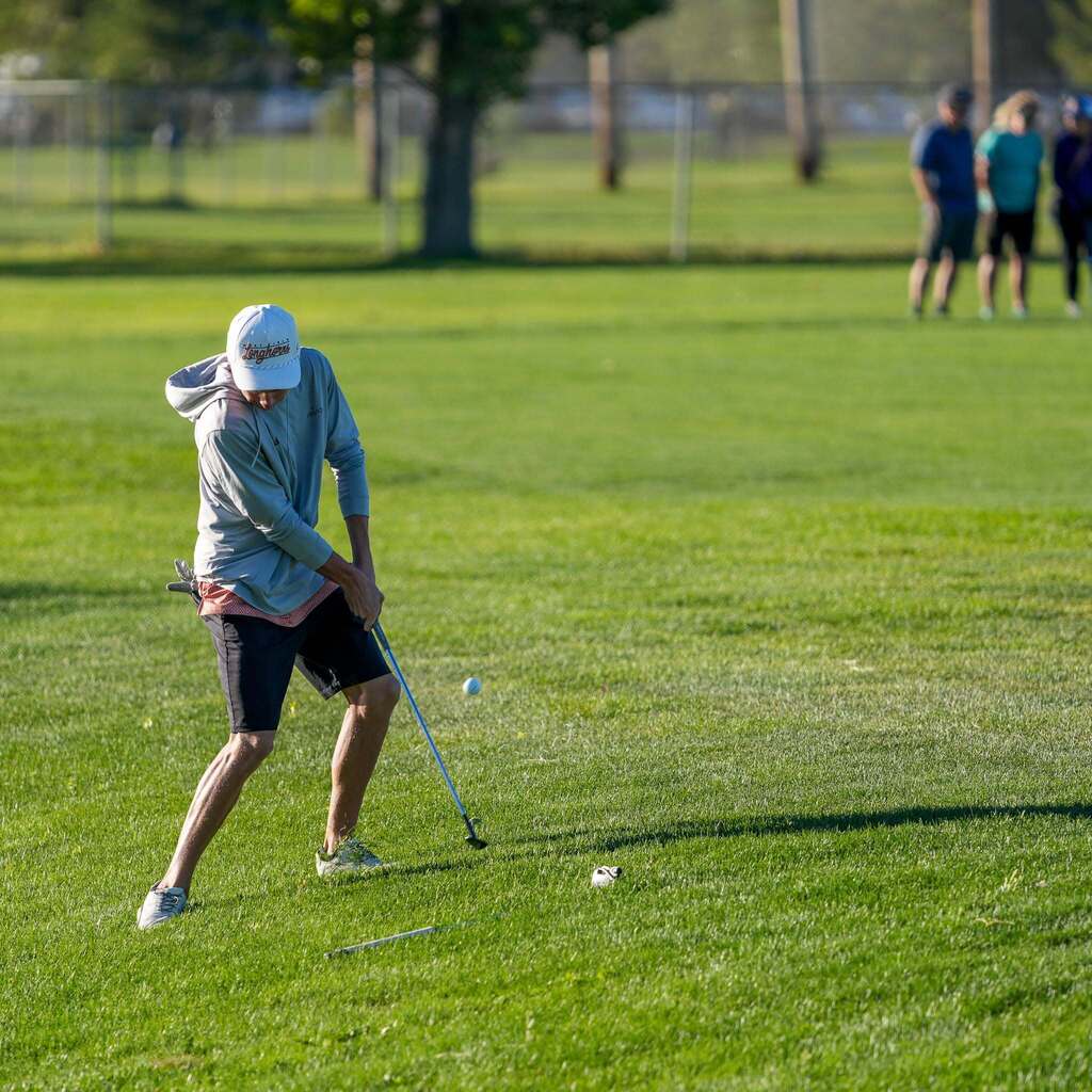 A male student-athlete wearing a grey Longhorns hoodie, white hat, and dark shorts watches his chip shot make contact with the golf ball on the fairway.