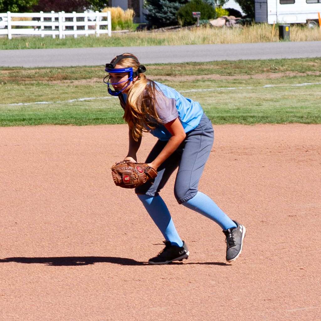 A Snowcrest softball infielder wearing a face mask and blue jersey stays low in an athletic ready stance, fielding her position on the dirt infield.