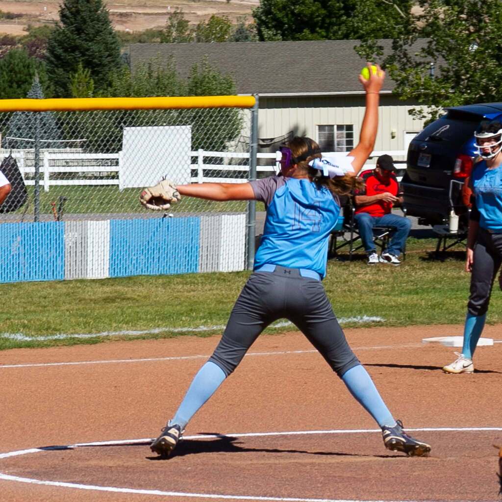 A Snowcrest Junior High softball pitcher in a blue jersey and protective face guard winds up and releases a pitch on the field during a game against Sand Ridge.