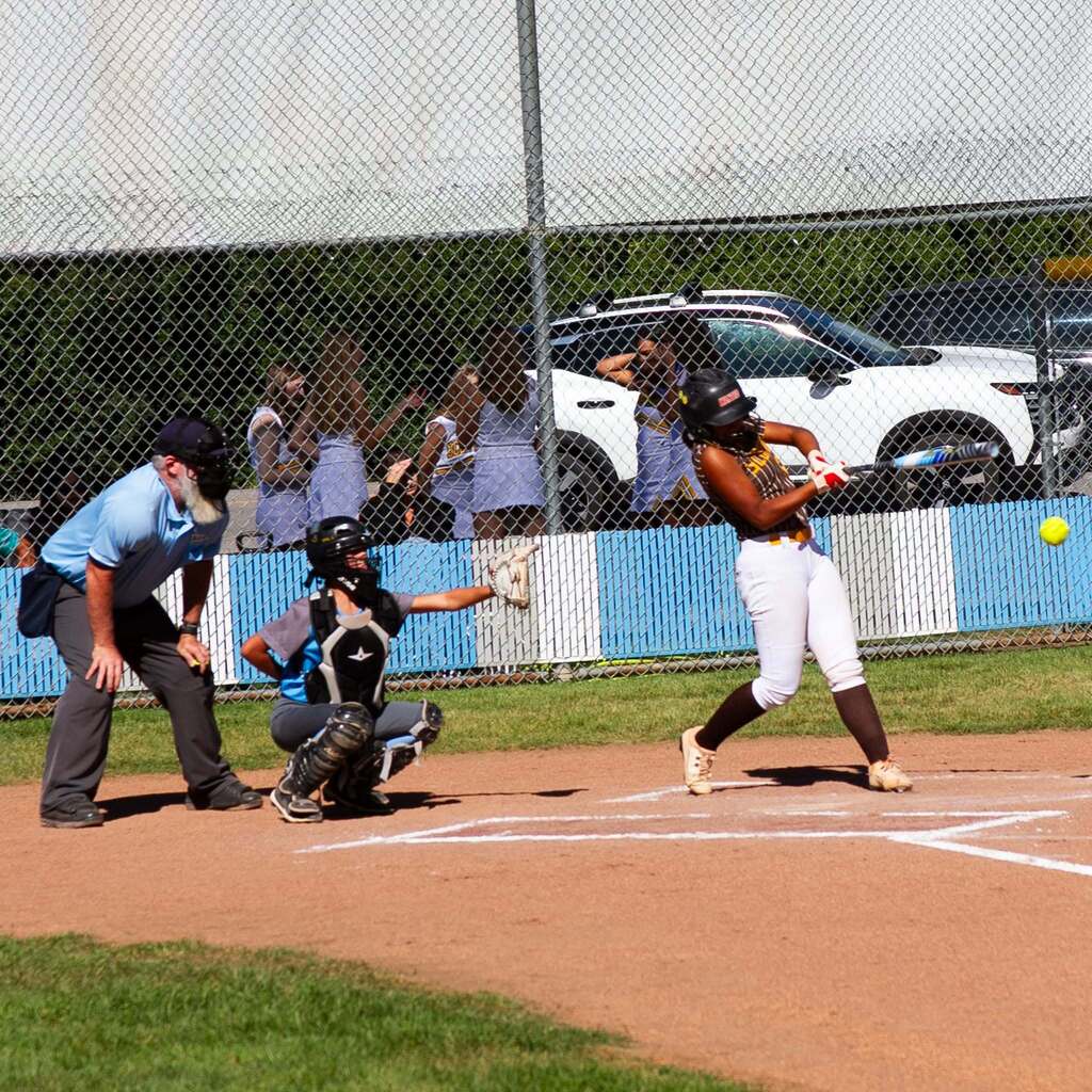 A Sand Ridge batter connects with a pitch, sending the softball flying into the field as the catcher and umpire track the ball.