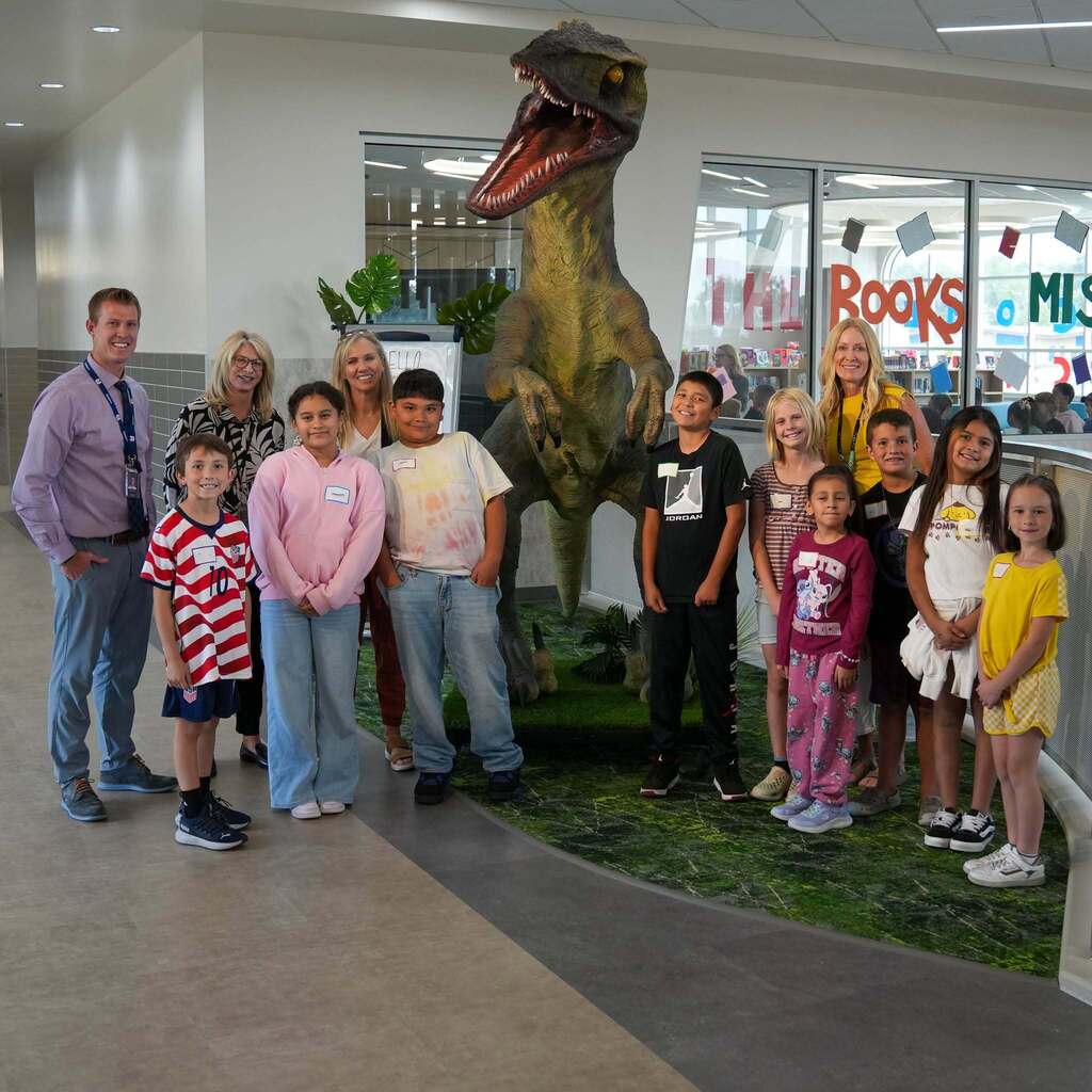 Representative Katy Hall, school administrators, and a group of Roosevelt Elementary student ambassadors pose for a group photo in front of a large life-sized dinosaur display inside the school.