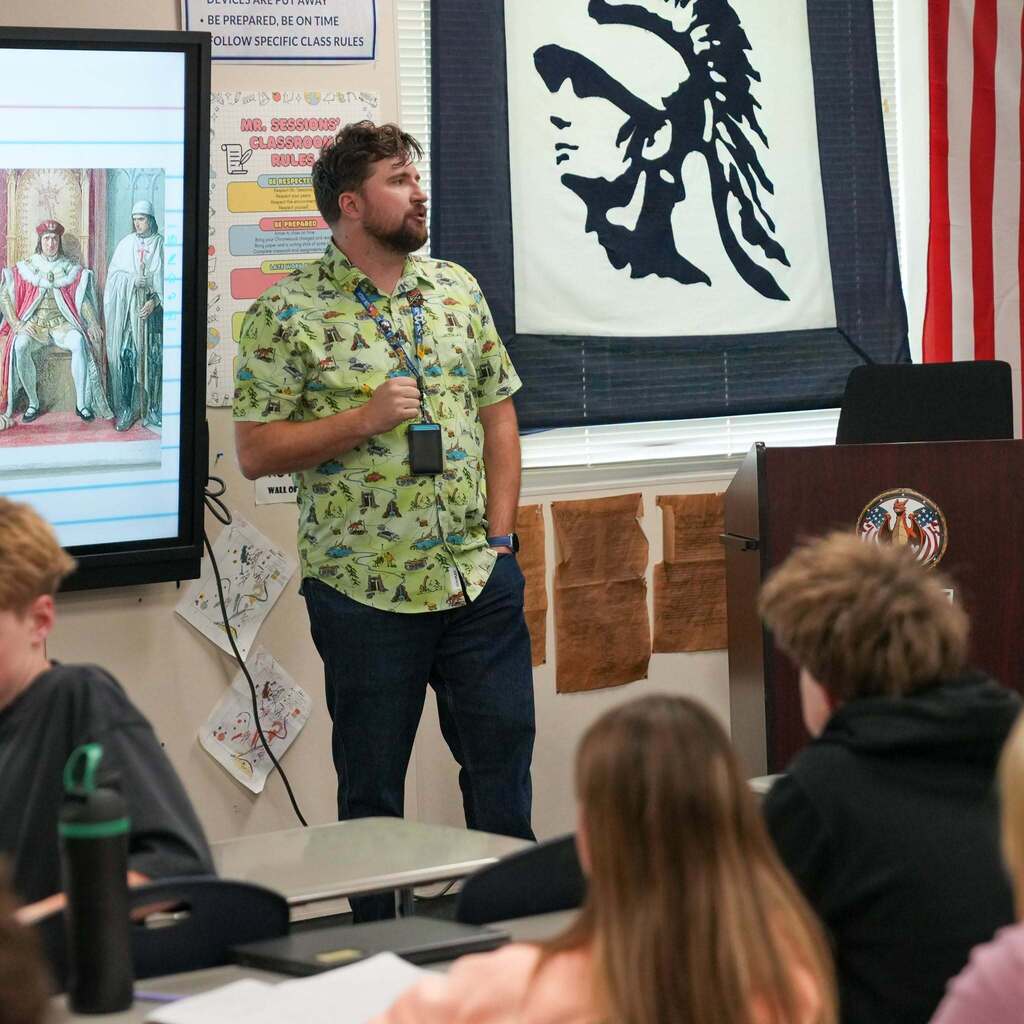 A teacher wearing a colorful patterned shirt addresses his class at the front of a South Ogden Junior High classroom next to a history presentation slide.