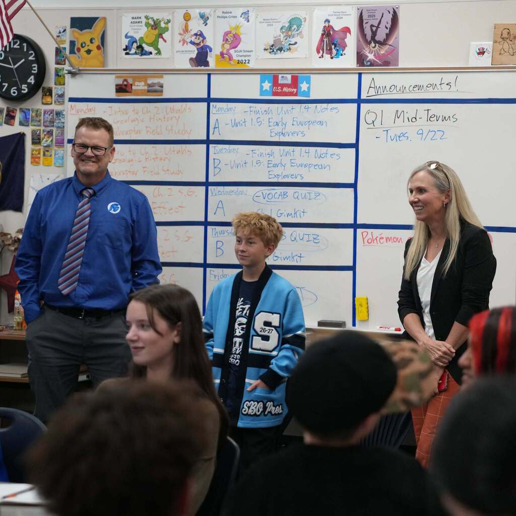 Representative Katy Hall and a South Ogden Junior High teacher smile while listening to an SBO student present in front of a whiteboard listing U.S. History class agendas.